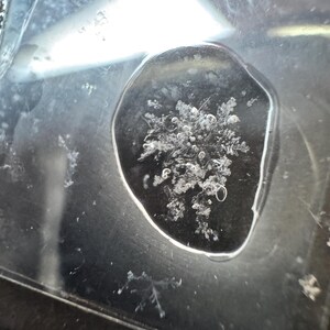 May include: A single, intricate white snowflake crystal is captured within a clear, circular droplet of water resting on a dark, reflective surface. The snowflake displays a complex, symmetrical, branching structure with delicate, icy details.
