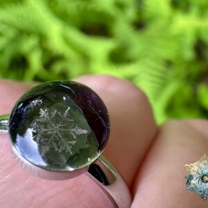 May include: A silver-toned ring featuring a smooth, rounded, dark-colored cabochon stone. The stone has a visible, intricate white snowflake pattern embedded within its surface. The ring band is polished metal and has a simple, clean design.