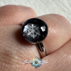 May include: A silver-toned ring featuring a circular, domed black cabochon with a white, intricate snowflake design embedded inside. The ring has a polished metal band and a smooth, reflective surface on the circular setting.