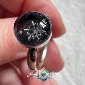 May include: A silver-toned ring featuring a circular, domed black resin setting that encapsulates a detailed white snowflake design. The text Snowflakes & Spiders is printed at the bottom with a small spider icon in the center of a snowflake.