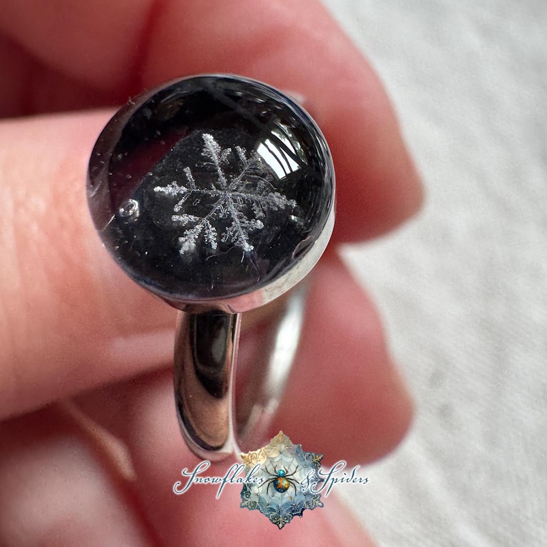 May include: A silver-toned ring featuring a circular, domed black resin setting that encapsulates a detailed white snowflake design. The text Snowflakes & Spiders is printed at the bottom with a small spider icon in the center of a snowflake.