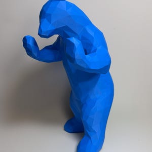 May include: A vibrant blue geometric bear sculpture. The bear is standing upright with its arms raised, showcasing a modern, angular design. The sculpture's surface is faceted, creating a play of light and shadow. This decorative piece adds a contemporary touch.