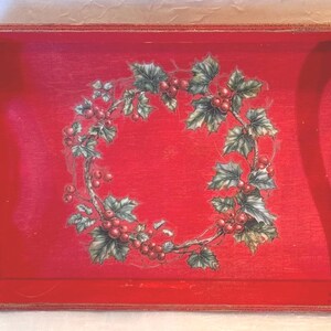 May include: A rectangular red wooden serving tray with curved handles. The tray features a wreath design of green leaves and red berries in the center, perfect for holiday decor.