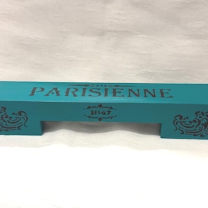 May include: A rectangular teal wooden riser with a cutout base. The top surface features the black text "PARISIENNE" and the number "1847" centered below it. Each side of the front face is decorated with a black ornate scrollwork pattern.