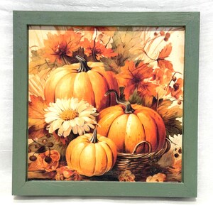 May include: A framed autumn-themed print featuring three orange pumpkins, a white daisy, and fall foliage. The pumpkins are arranged in front of a backdrop of orange and yellow leaves. The frame is a muted green color.