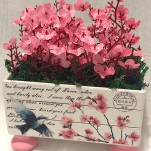 May include: A rectangular white wooden planter box featuring pink cherry blossom branches, a blue bird illustration, and cursive text. The box is filled with artificial pink flowers and green moss, supported by four small, round, light pink wooden feet.