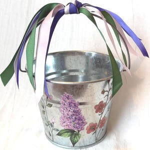 Könnte beinhalten: Ein verzinkter Metalleimer mit einem Blumenmuster aus violetten Fliederblüten und kleinen rosa Blüten. Der Eimer hat einen Metallgriff, der mit mehreren oben zusammengebundenen Bändern in den Farben Helllila, Dunkellila und Waldgrün verziert ist.