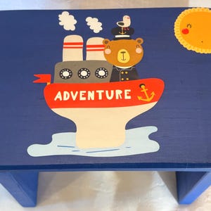 Puede incluir: Un taburete azul de madera con temática náutica. El taburete presenta un oso de dibujos animados capitán en un barco con la palabra "ADVENTURE" en el lateral. También se representa un sol sonriente y una gaviota.