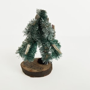 May include: A small, artificial Christmas tree with green and white accents. The tree is made of bristled material and is mounted on a round, dark brown wooden base. The tree has a rustic, vintage aesthetic.