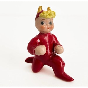 May include: A small, vintage figurine of a kneeling figure in a red jumpsuit. The figurine has a fair complexion, rosy cheeks, and yellow hair. It has small golden horns and is posed on a white surface.