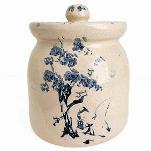 May include: A cream-colored ceramic jar with a matching lid, featuring a speckled finish and a hand-painted blue design of a tree and a figure fishing. The jar has a cylindrical body with a slightly rounded shoulder and a knobbed lid handle.