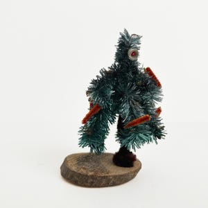 May include: A small, handmade Christmas tree with dark green needles and brown pipe cleaner accents. The tree is adorned with a red jewel and sits on a wooden base, evoking a vintage aesthetic.