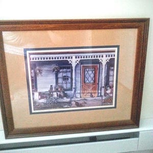 May include: Framed print of a quaint cottage porch scene. The artwork features a wooden door with a diamond pattern, a window, and a small table with plants. The frame is a dark brown color.