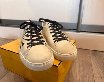 May include: A pair of cream-colored sneakers with black laces, resting on a yellow box. The sneakers have a low-top design with a white sole and a subtle pattern on the side. The laces are neatly tied.