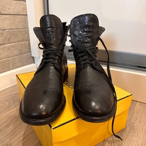 Könnte beinhalten: Ein Paar schwarze Leder-Schnürstiefel mit strukturierter Oberfläche. Die Stiefel werden auf einer gelben Schachtel präsentiert. Die Stiefel haben eine runde Spitze und eine robuste Sohle. Die Schnürsenkel sind schwarz und gebunden.