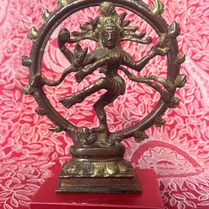 May include: A gold-toned metal statue of the Hindu deity Shiva in the Nataraja pose, dancing on a demon. The statue is on a red box.