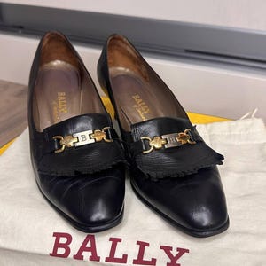May include: A pair of black leather Bally loafers with gold-tone hardware and fringe detailing. The shoes are displayed on a white cloth bag with the Bally logo in red. The shoes have a low heel.