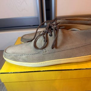 May include: Light brown suede ankle boots with brown laces, on a yellow box. The boots have a rounded toe and a light yellow sole. The laces are tied. The boots are approximately 25 cm long.
