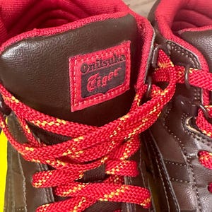 May include: Close-up of brown and red high-top trainers. The trainers have red laces with yellow detailing, a red interior, and a brown exterior with the "Onitsuka Tiger" logo on the side.