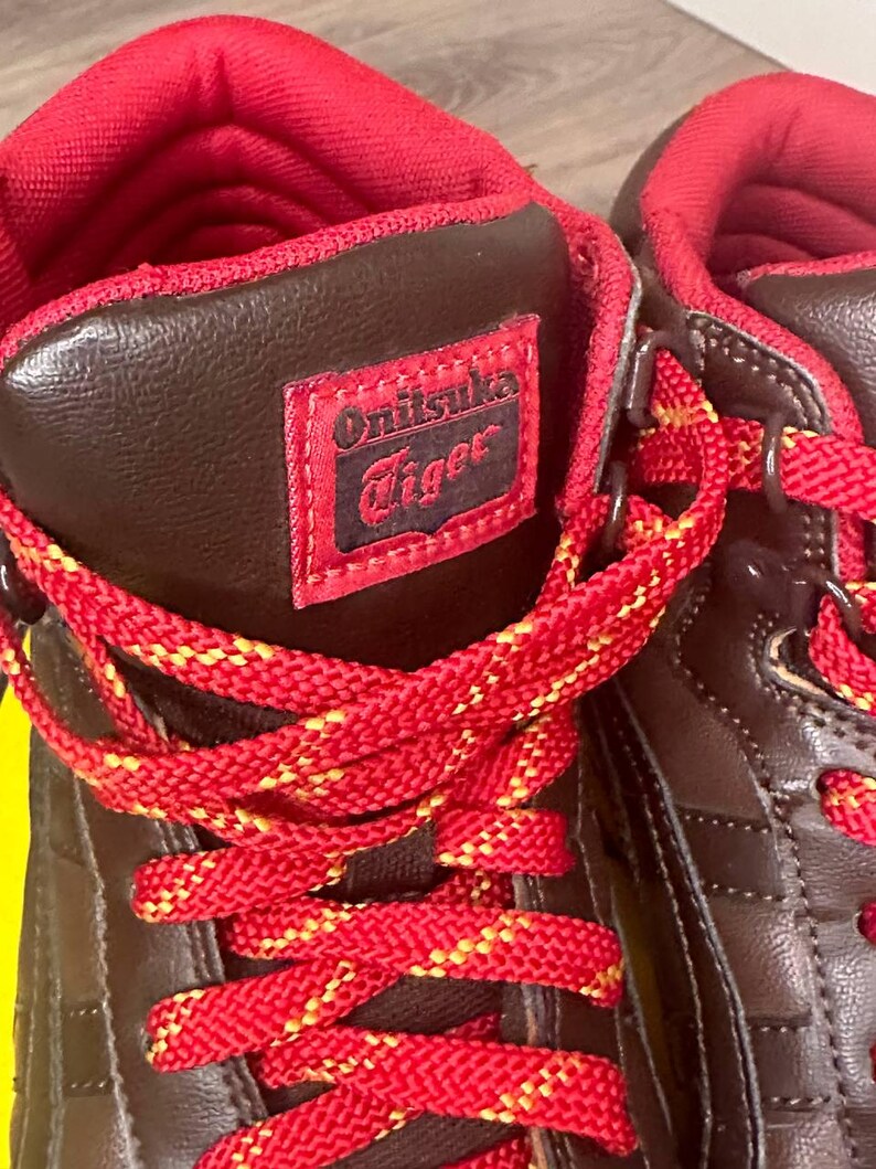 May include: Close-up of brown and red high-top trainers. The trainers have red laces with yellow detailing, a red interior, and a brown exterior with the "Onitsuka Tiger" logo on the side.