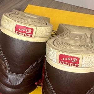 May include: A pair of dark brown high-top trainers featuring a textured off-white rubber sole with a red rectangular patch on the heel displaying the white text Onitsuka Tiger. The shoes have a smooth leather upper and red laces.