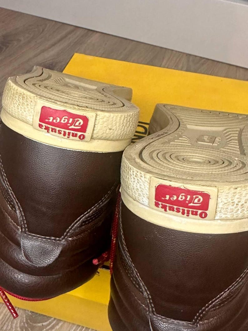May include: A pair of dark brown high-top trainers featuring a textured off-white rubber sole with a red rectangular patch on the heel displaying the white text Onitsuka Tiger. The shoes have a smooth leather upper and red laces.