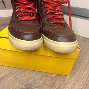 May include: Brown high-top trainers with red and gold laces, resting on a yellow box. The trainers feature a cream sole and the brand name "Onitsuka Tiger" is visible on the tongue.