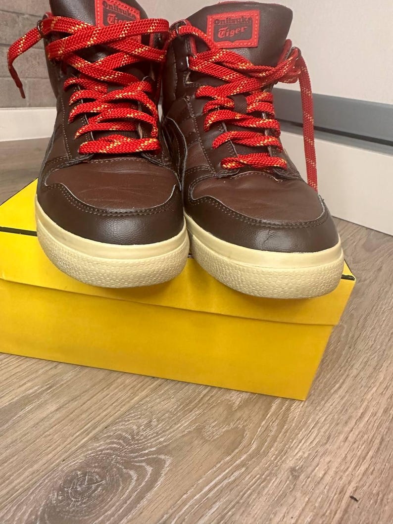 May include: Brown high-top trainers with red and gold laces, resting on a yellow box. The trainers feature a cream sole and the brand name "Onitsuka Tiger" is visible on the tongue.