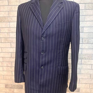 May include: A navy blue pinstripe blazer with a three-button fastening. The jacket features a notched lapel, a chest pocket, and two front pockets. White pinstripes run vertically. The blazer is displayed on a black mannequin against a brick wall.