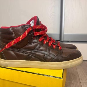 May include: Brown high-top trainers with red laces. The shoes have a brown leather upper with a quilted design and a cream-coloured sole. The interior is red. The laces are red with yellow accents. The trainers are on a yellow box.