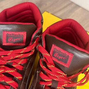 May include: Close-up of two brown and red high-top trainers. The trainers have red laces with yellow accents and the brand name "Onitsuka Tiger" on a red patch. The interior lining is red.