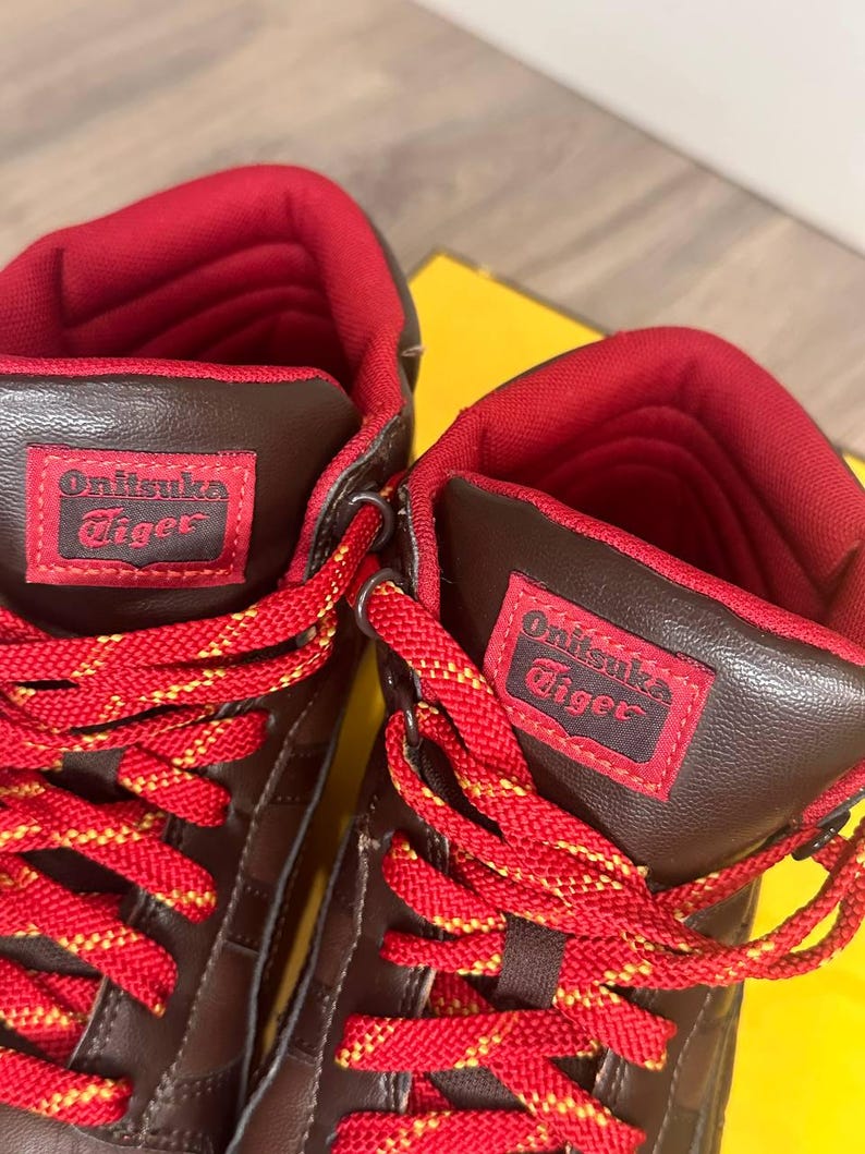 May include: Close-up of two brown and red high-top trainers. The trainers have red laces with yellow accents and the brand name "Onitsuka Tiger" on a red patch. The interior lining is red.