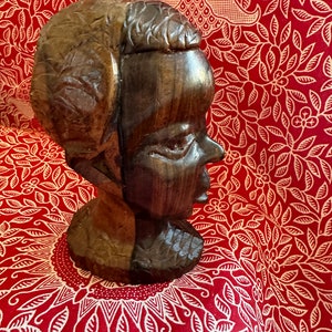 May include: A dark brown wooden sculpture of a person's head and shoulders. The sculpture is carved in a realistic style and has a detailed face.