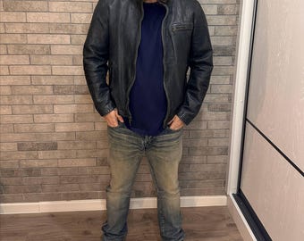 May include: A black leather jacket with a zip, worn over a navy blue t-shirt and faded blue jeans. The outfit is completed with black and white trainers. The person is standing in front of a brick wall.