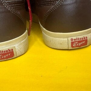 May include: A pair of brown leather trainers with white soles and the "Onitsuka Tiger" logo in red. Red laces are visible. The shoes are on a yellow surface.