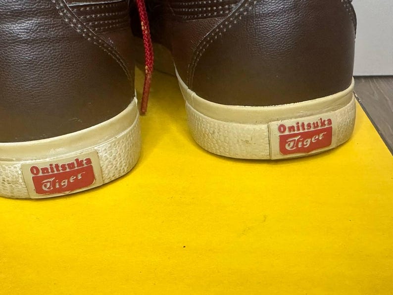 May include: A pair of brown leather trainers with white soles and the "Onitsuka Tiger" logo in red. Red laces are visible. The shoes are on a yellow surface.