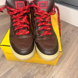 May include: Brown high-top trainers with red and yellow laces. The trainers have a brown leather upper and a cream-coloured sole. The tongue of the shoe has the text "Onitsuka Tiger" in white.
