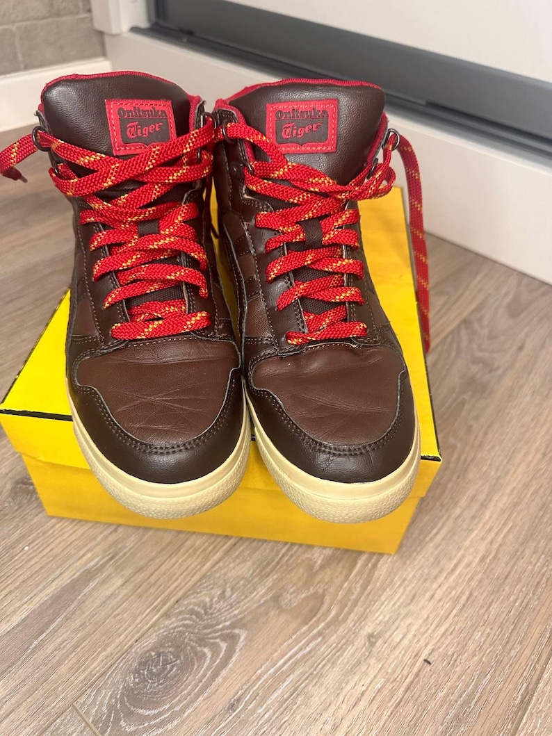 May include: Brown high-top trainers with red and yellow laces. The trainers have a brown leather upper and a cream-coloured sole. The tongue of the shoe has the text "Onitsuka Tiger" in white.