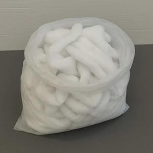 May include: A clear plastic bag filled with fluffy, white polyester fiberfill. The bag is open, revealing the soft, cloud-like material inside. The bag is sitting on a gray surface.