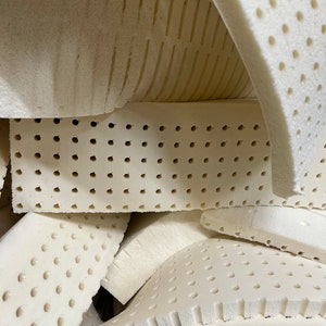 May include: A close-up of a pile of white latex foam mattress toppers with a variety of hole patterns. The foam is cut into different shapes and sizes.