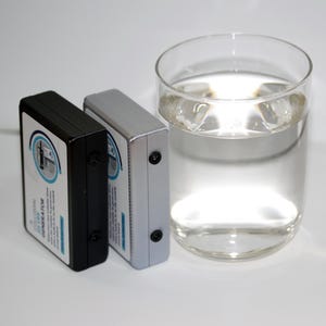 May include: Two rectangular devices, one black and one silver, are positioned next to a clear glass of water. The devices have a label with text and a circular design. The glass is filled with clear liquid and ice cubes.