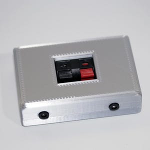 May include: A silver rectangular electronic component with a central recessed area featuring black and red terminals. Two small black circular ports are visible on the side. The component has a textured surface.