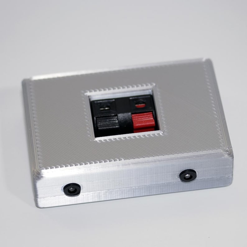 May include: A silver rectangular electronic component with a central recessed area featuring black and red terminals. Two small black circular ports are visible on the side. The component has a textured surface.