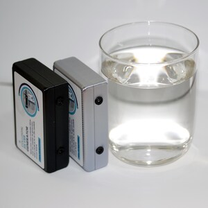 May include: Two rectangular devices, one black and one silver, are placed beside a clear glass of water. The devices have a label with text and a circular design. The glass contains clear liquid and ice cubes.