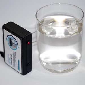 May include: A black device with a white label and the text "Colloidal Silver Generator" next to a clear glass of water. The device has a red light and a power cord. The glass is filled with clear liquid.