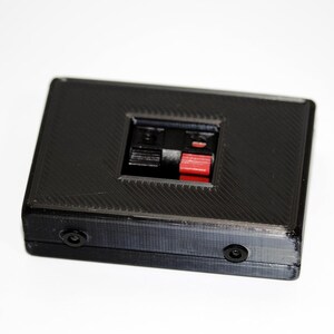 May include: A black rectangular electronic device with a textured surface. It features a central recessed area with black and red connectors, and two small circular ports on the side. The device is likely used for audio or electronic connections.