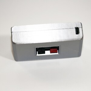 May include: A rectangular, silver-coloured electronic device with a black and red port. The device has a small, rectangular opening on one side and a small black port on the top. Likely used for audio or video connections.