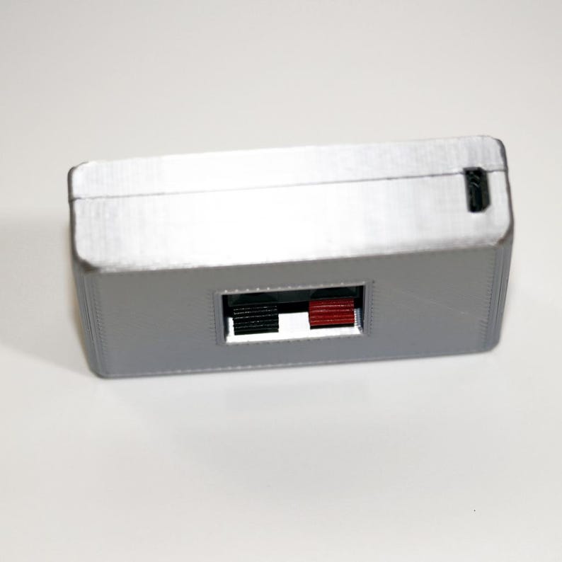 May include: A rectangular, silver-coloured electronic device with a black and red port. The device has a small, rectangular opening on one side and a small black port on the top. Likely used for audio or video connections.