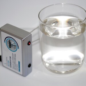 May include: A silver colloidal silver generator with a red light and a glass of clear liquid. The generator has a label with blue and white text and a circular graphic. A black cord is connected to the generator.