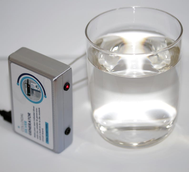 May include: A silver colloidal silver generator with a red light and a glass of clear liquid. The generator has a label with blue and white text and a circular graphic. A black cord is connected to the generator.